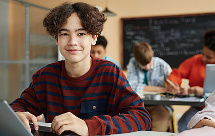 Schüler mit lockigem, braunen Haar lächelt in die Kamera. Er sitzt im Klassenzimmer und arbeitet am Laptop.