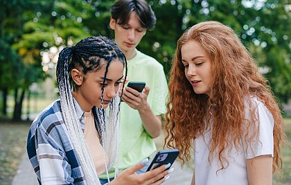 Zwei Teenager-Mädchen und ein Teenager-Junge schauen auf ihre Handys.