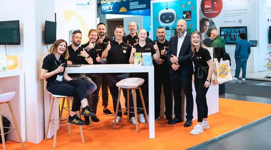 LMZ Team Man sieht eine Gruppe von Mitarbeitenden des Landesmedienzentrums, die in die Kamera lachen. Sie befinden sich am Messestand des LMZ.