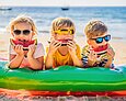 Sharenting – Kinderbilder teilen, aber wie? Drei Kinder im Grundschulalter liegen bäuchlings auf einer Luftmatratze am Strand. Ihre Gesichter sind durch Sonnenbrillen und Melonenschnitze vor dem Mund teilweise verdeckt.