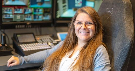 Barbara Glück vom LMZ Studio Eine junge Frau mit hellbraunen langen Haaren und runder Brille sitzt auf einem Regiestuhl. Hinter ihr sind das Regiepult und Monitore zu sehen.