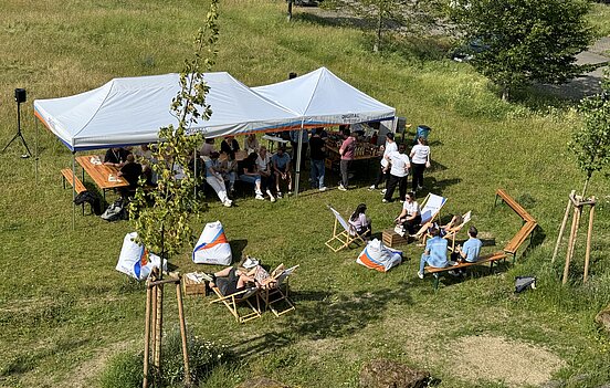 Das Bild zeigt eine Outdoor-Veranstaltung auf einer Wiese bei sonnigem Wetter. In der Mitte des Bildes steht ein großes weißes Zelt, unter dem mehrere Menschen auf Bänken und an Tischen sitzen. Im Vordergrund des Bildes sind mehrere Liegestühle und Sitzsäcke verteilt, auf denen Menschen entspannt sitzen und sich unterhalten.