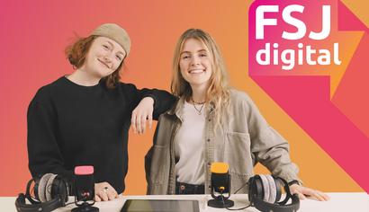 FSJ digital Zwei junge Frauen stehen lächelnd vor einem Tisch mit zwei Mikrofonen, Kopfhörern und einem Tablet.