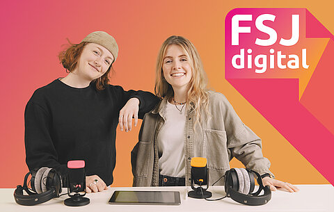 FSJ digital Zwei junge Frauen stehen lächelnd vor einem Tisch mit zwei Mikrofonen, Kopfhörern und einem Tablet.