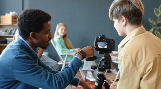 Drehort Klassenzimmer Ein Mann zeigt einer Teenagerin etwas auf einer Filmkamera, die auf ein weiteres Mädchen im Hintergrund gerichtet ist.