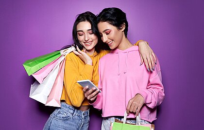 Zwei Teenager-Mädchen in pastellfarbenen Hoodies und mit Einkaufstüten beladen schauen lachend auf ein Smartphone.