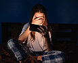 Mädchen sitzt im Dunklen auf dem Bett und schaut zwischen den Fingern auf ihr Smartphone.