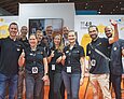 LEARNTEC-Team des LMZ 2024 Zehn Frauen und Männer in schwarzen Polo-T-Shirts mit dem Logo des Landesmedienzentrums zeigen lachend "Daumen hoch" in die Kamera. Im Hintergrund ist ein weiß-orange-farbener Messestand zu sehen.