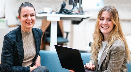 Karriere am LMZ Man sieht zwei junge Frauen, die laechelnd in die Kamera schauen. Die Frau links hat dunkelbraune Haare, die Frau rechts haelt einen Laptop in der Hand und hat dunkelblonde Haare. Sie sitzen auf einem Sofa im Buero des Landesmedienzentrums.