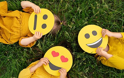 Drei Kinder in gelben Oberteilen liegen auf einer grünen Wiese und halten Emojis aus Pappkarton vor ihre Gesichter.