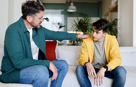 Vater und Sohn im ernsten Gespräch über sexuelle Belästigung Ein Vater legt seinem Teenager-Sohn verständnisvoll die Hand auf die Schulter.