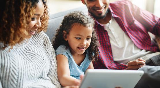 Eltern-Medienmentoren-Programm Ein Kind sitzt zwischen seiner Mutter und seinem Vater auf dem Sofa. Alle schauen gemeinsam in ein Tablet.