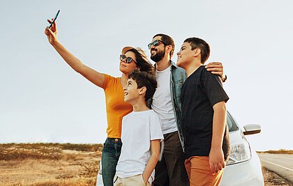 Eine Frau hält ein Handy in die Höhe, um ein Selfie mit ihrem Mann und ihren zwei Söhnen zu machen. Sie stehen vor einem weißen Auto in einer trockenen Steppenlandschaft.