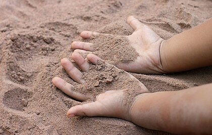 Zwei Kinderhände liegen mit dem Handteller nach oben im Sand und sind mit diesem bedeckt..