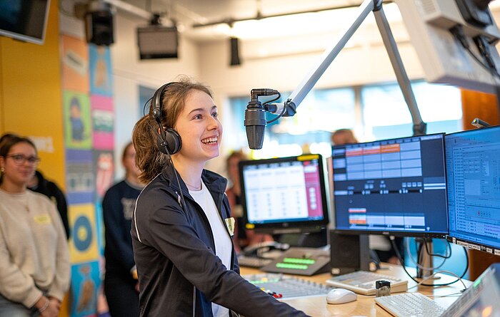 Schüler-Medienmentoren-Programm Radioworkshop Ferienkurs Ein Maedchen mit dunkelblonden Haaren hat einen Kopfhoerer auf und spricht in ein Mikrofon. Sie steht in einem Radiostudio und lacht.